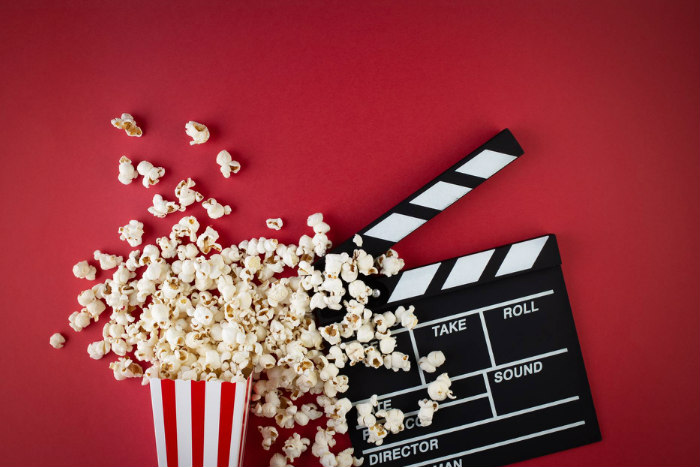 Film reel and a spilled container of popcorn against a red background 