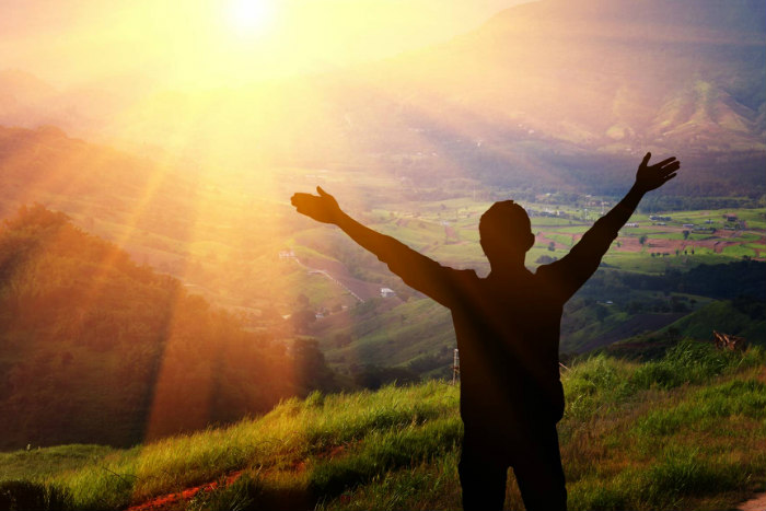 Man facing away from the camera with his arms raised as he looks out from a mountaintop 