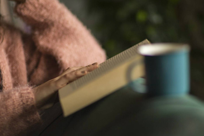 Closeup of a hand placed on a book as if reading while curled up beneath a blanket near a mug 