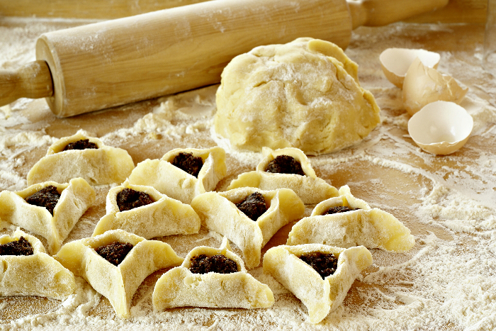 Hamantaschen filled with a dark filled next to an unshaped ball of dough and a rolling pin