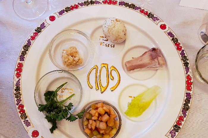 Closeup of a white seder plate with a colorful rim set upon a white tablecloth 