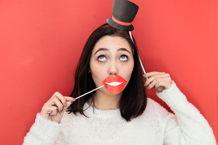 Young brunette woman holding lips and a tiny hat on sticks up to her face as if in a photo booth