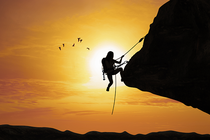 man hanging off the side of a mountain with the sun behind him
