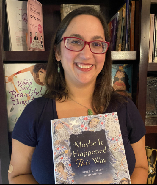 A woman with glasses and straight brown hair stands smiling in front of a bookcase holding a book titled "Maybe It Happened This Way"