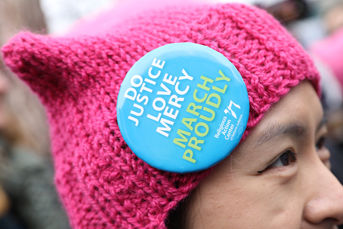 an image of a woman wearing a pink winter hat with a pin on it that says "Do Justice Love Mercy March Proudly" with a white RAC logo