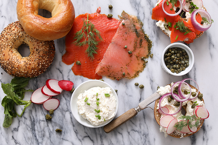 multiple bagels, two full and one sliced in half, with lox, cream cheese and a bowl of capers on a white and gray marble countertop