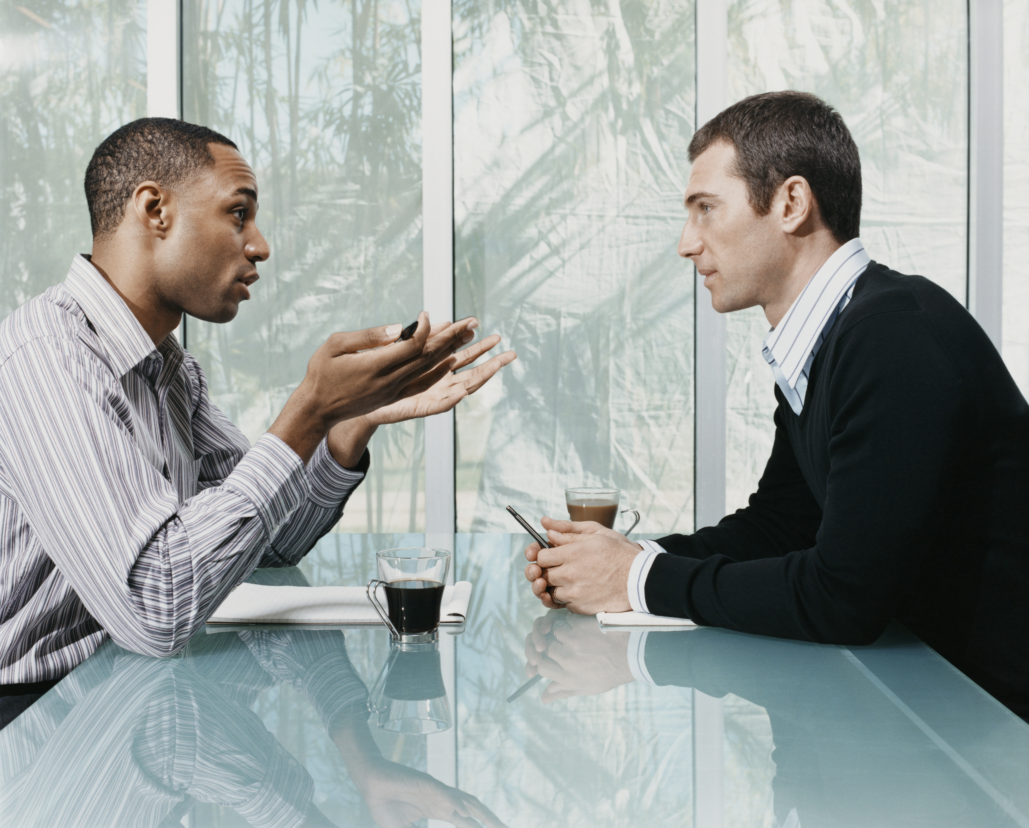 Photo of two men talking, sitting at a table with coffee in front of them