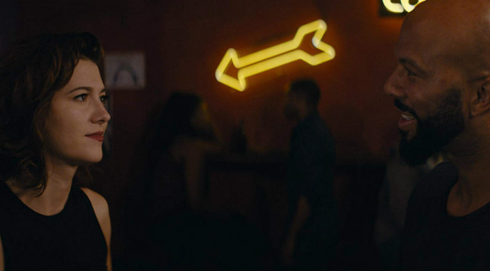 Movie still of a white woman speaking with a black man with a neon arrow shining between as at a bar