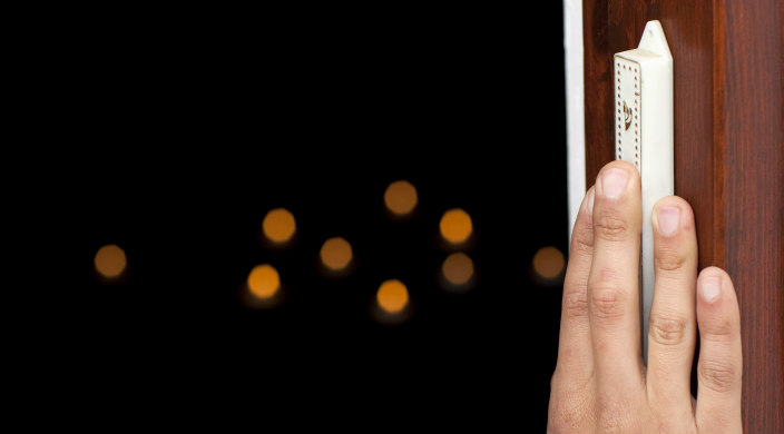 A hand lightly touches a mezuzah on a doorpost with black space behind the entryway