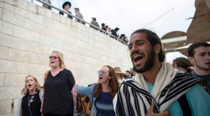 The author wears a prayer shawl and sings as he walks with other Women of the Wall members toward the Kotel while Haredi look on from atop a tall wall