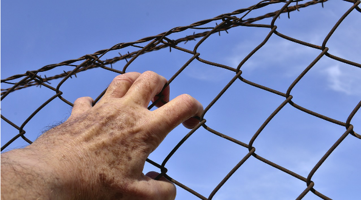 A mans hand clutching at a barbed wire fence against a blue sky 