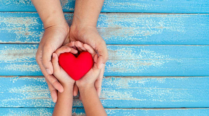 Adult hands cupping a childs hands holding a red plastic heart atop a blue wooden table