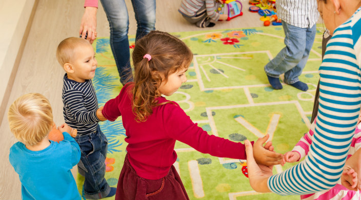 Young children in a classroom setting dancing or moving in a circle while holding hands with one another and their adult instructors