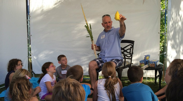 Rabbi Paul Kipnes hold a lulav and an etrog while standing under a sukkah and teaching a group of young children 