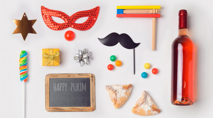 Aerial flatlay of a variety of Purim related objects and items including masks hamantaschen and a bottle of wine 