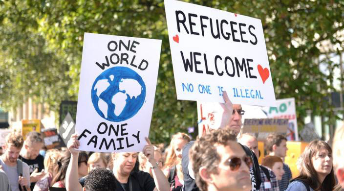 Protesters holding signs that read ONE WORLD ONE FAMILY and REFUGEES WELCOME NO ONE IS ILLEGAL
