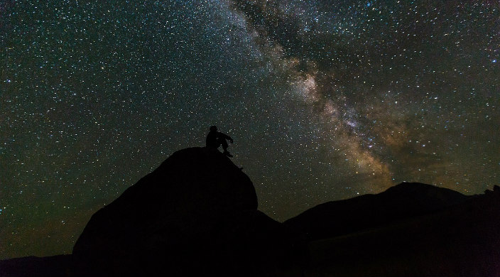 Silhouette of a man sitting on rocks looking out at a starry night sky 