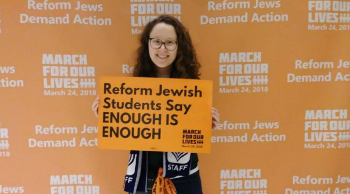 Jewish young adult holding a sign about gun violence prevention in front of a March for Our Lives banner
