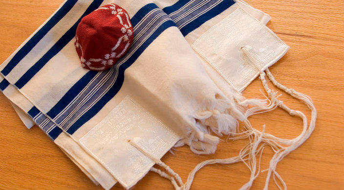 red knit kippah lying atop a blue and white fringed prayer shawl