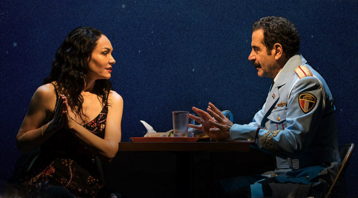 A still from the Broadway show The Bands Visit with a young woman speaking across a table to an older man in uniform