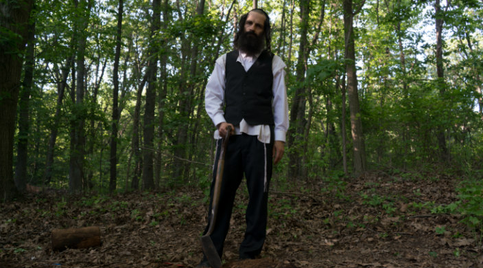 Movie still of an Orthodox Jewish man holding a shovel and standing atop a mound of dirt as if digging a grave