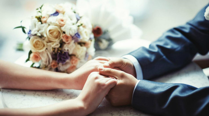 Closeup of a bride and groom holding hands with a bouquet of wedding flowers on the table next to them 