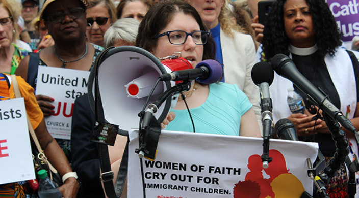 Female speakers in front of a microphone and a podium that reads WOMEN OF FAITH CRY OUT FOR IMMIGRANT CHILDREN