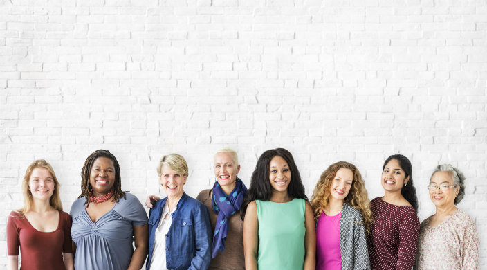 Smiling women of all ages and ethnicities wearing brightly colored clothing and standing in a line against a white brick wall