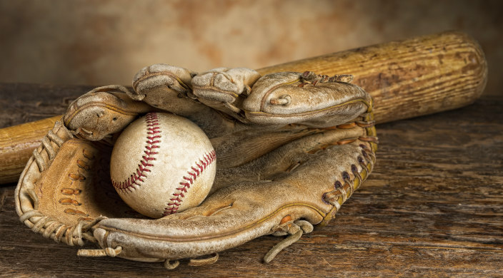 worn baseball glove holding baseball in foreground; wooden baseball bat in back