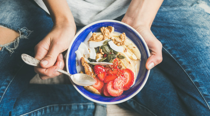 Overhead shot of a person sitting cross-legged on the floor holding a bowl of granola, seeds, and fruit