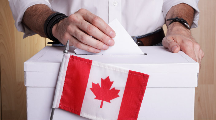 A mans hands dropping a ballet into a ballot box with a Canadian flag in front