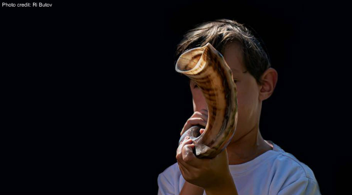 Young child blowing a shofar against a black background