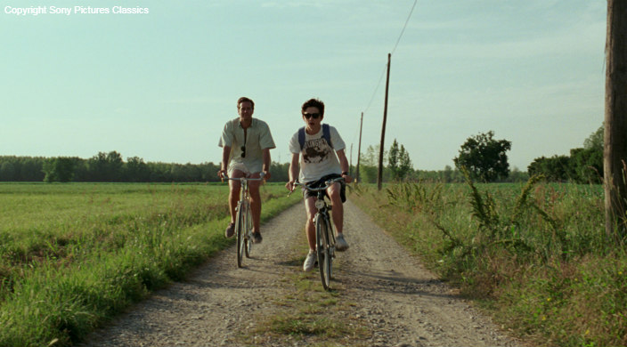 Screenshot from the movie Call Me By Your Name shows two young men riding bicycles down a dirt path 