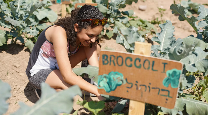 Smiling teen girl digging in the dirt next to a hand painted sign that says BROCCOLI in both Hebrew and English 