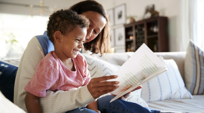 Mom holds a laughing son on her lap as she reads a book to him from the couch