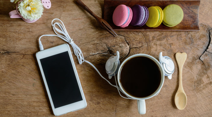 Cell phone with earbuds, coffee, wooden spoon with a heart handle and four pastel macaroons all seen from above