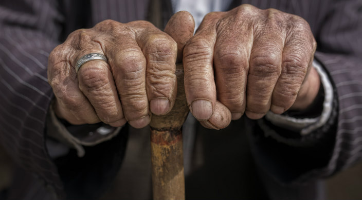 Leathery hands of a man in a shirt and suit jacket clutching the top of a weathered, wooden cane