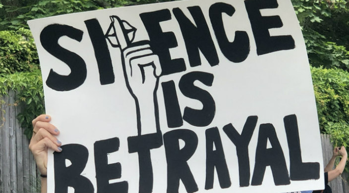 White hands holding a sign that reads SILENCE IS BETRAYAL