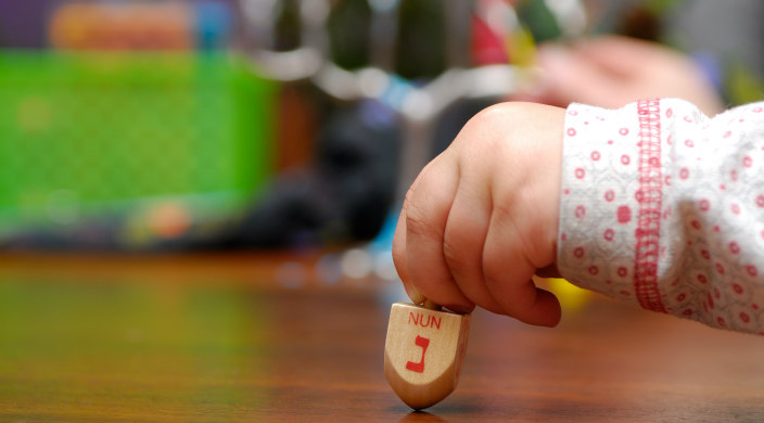 A toddler's hand holding a dreidel in the foreground; hanukkiyah and gifts in the background (blurred)