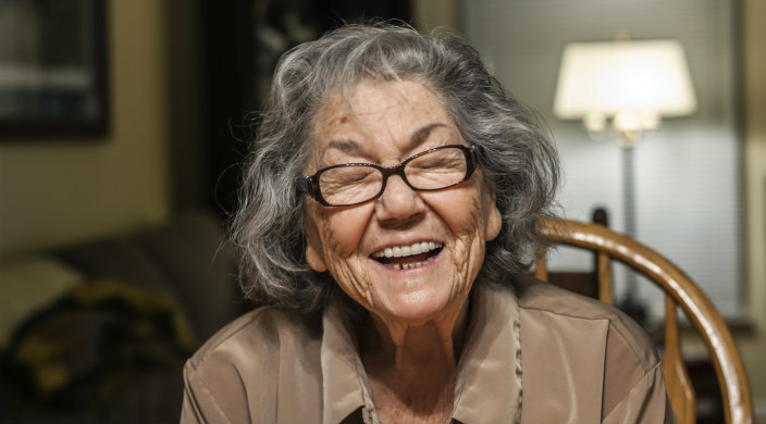 Older woman laughing toward the camera while sitting at a kitchen table