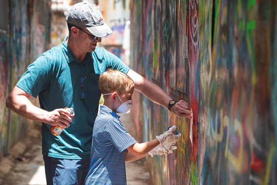 an image of a father and son standing next to a wall full of paint, holding spray paint cans