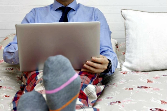 Man holding a laptop in bed while wearing a shirt and tie on top with plaid pajama pants on the bottom
