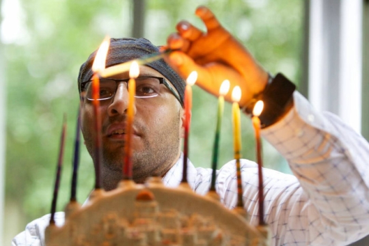 Closeup of a man in a kippah lighting a full Hanukkah menorah