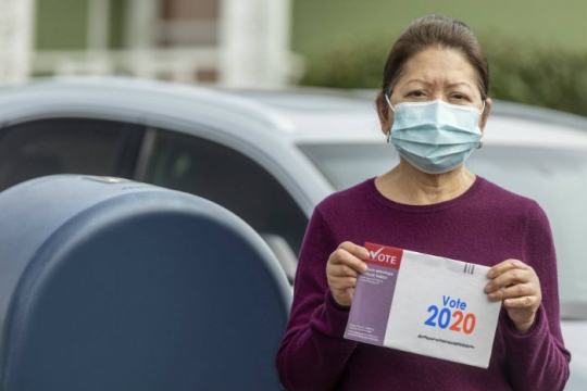 Woman wearing a face mask and holding a mail in ballot while standing next to a USPS mailbox