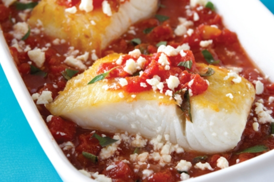 Cuts of a white fish in a pan with tomato sauce white cheese and green herbs