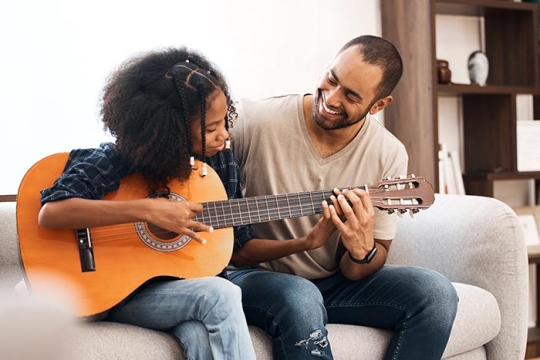 Parent sitting with child and smiling while child plays guitar