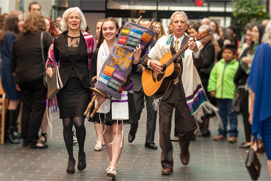 A teenager holding a Torah next to a woman and a man playing guitar, all walking through a synagogue