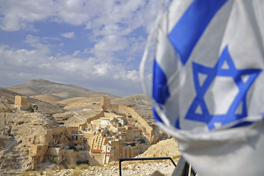 an image of an Israeli flag with small town and mountains in the background.