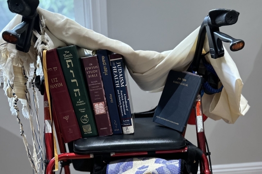 A red walker with a tallit draped over the handlebars and a Torah, siddur, Jewish Study bible, copy of Etz Hayim, and Rabbi's manual on the seat