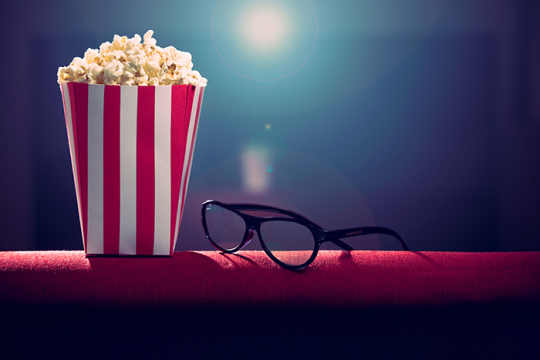 an image of a pair of black glasses sitting next to movie theater popcorn on a red chair with a light shining bright behind